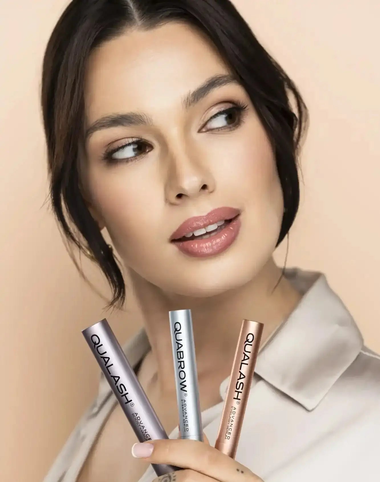 The model holds three sleek, metallic cosmetic applicators—two in silver and one in rose gold—with the brand name ’qualash’ prominently displayed on each.